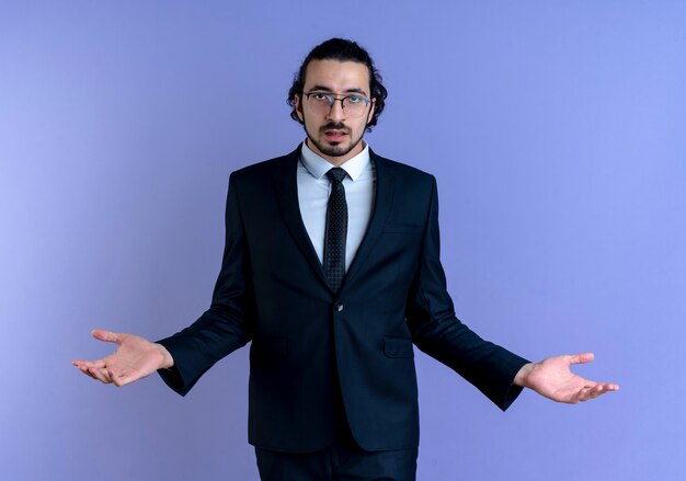 Business man in black suit and glasses looking confused and uncertain spreading arms to the sides standing over blue wall