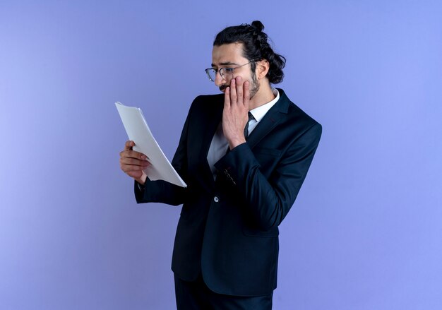 검은 양복과 파란색 벽 위에 서있는 그의 얼굴에 손으로 걱정 문서를보고 안경 비즈니스 남자