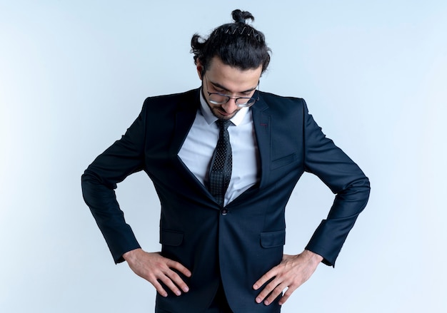 Business man in black suit and glasses looking down with arms at hip tired and bored standing over white wall