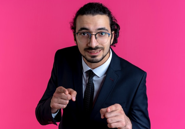 Business man in black suit and glasses looking to the front clenching fists happy and excited standing over pink wall