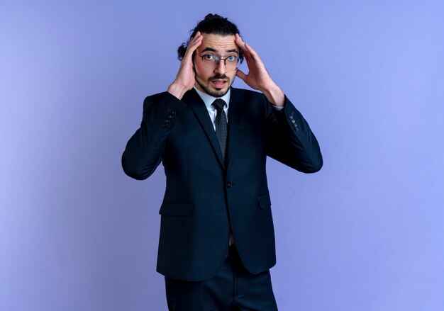 Business man in black suit and glasses looking to the front confused with hands near head standing over blue wall