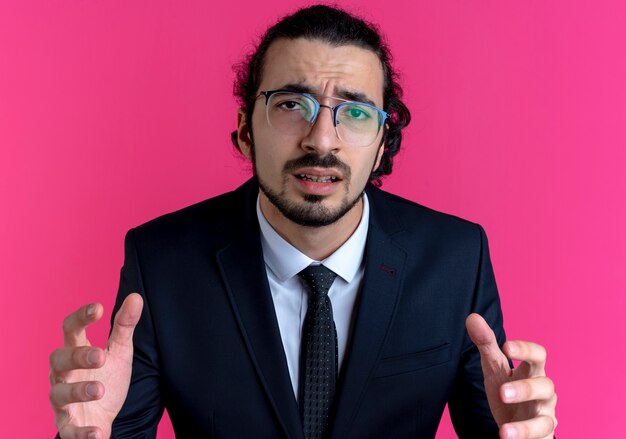 Business man in black suit and glasses looking to the front confused with raised arms standing over pink wall