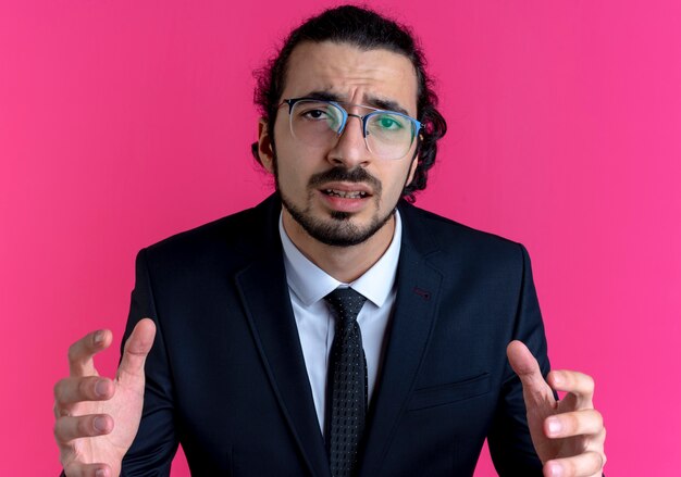 Деловой человек в черном костюме и очках смотрит вперед, смущенный поднятыми руками, стоя над розовой стеной