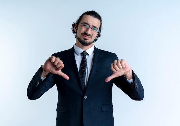 Business man in black suit and glasses looking to the front displeased showing thumbs down standing over white wall