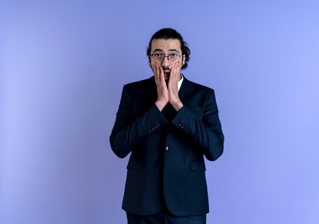 Business man in black suit and glasses looking to the front shocked covering mouth with hand standing over blue wall