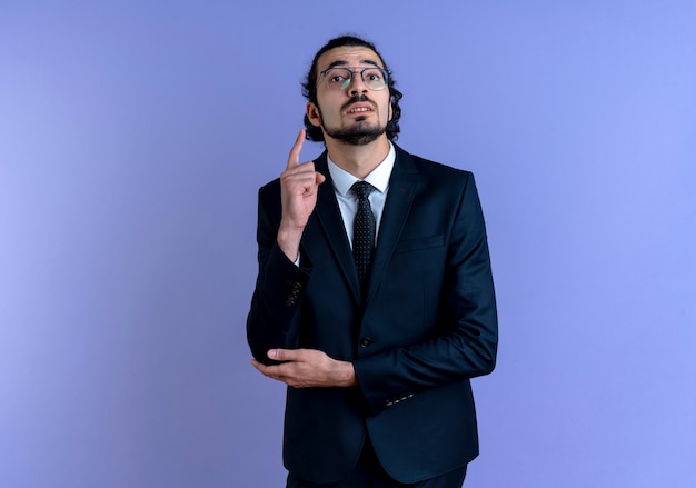 Business man in black suit and glasses looking to the front showing index finger reminding himself not to forget standing over blue wall