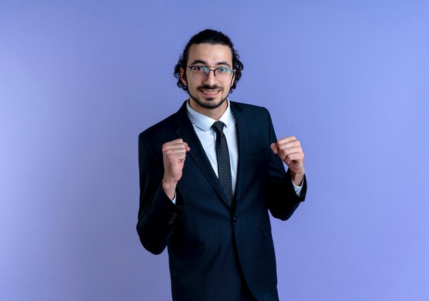 Business man in black suit and glasses looking to the front smiling cheerfully clenching fists happy and excited standing over blue wall