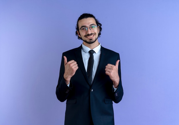 Business man in black suit and glasses looking to the front smiling cheerfully showing thumbs up standing over blue wall