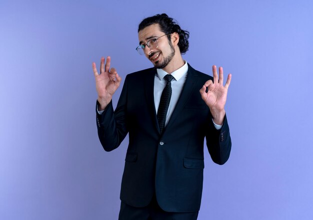 Деловой человек в черном костюме и очках смотрит вперед, улыбаясь, делая знак ОК, обеими руками стоя над синей стеной