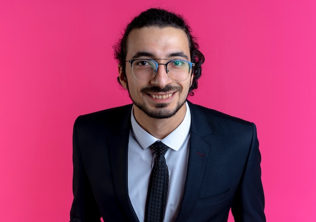 Business man in black suit and glasses looking to the front smiling with happy face standing over pink wall