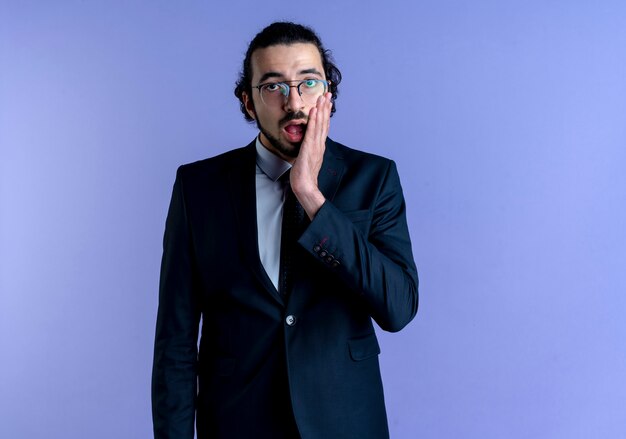 Business man in black suit and glasses looking to the front surprised and confused standing over blue wall