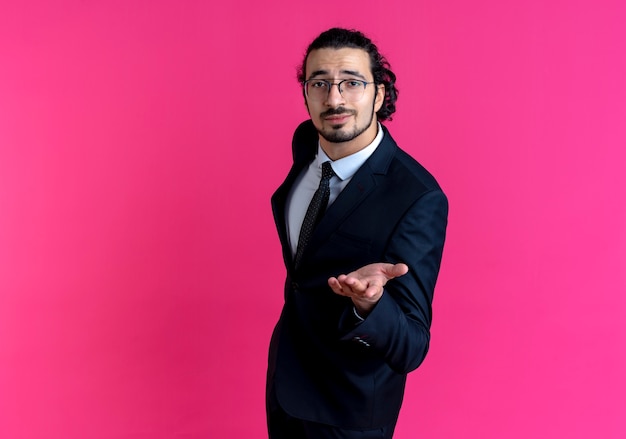 Business man in black suit and glasses looking to the front with arm out as asking question standing over pink wall