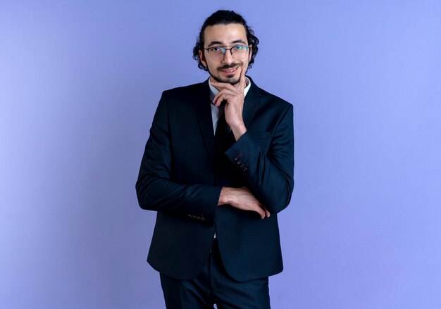 Business man in black suit and glasses looking to the front with hand on chin smiling confident standing over blue wall