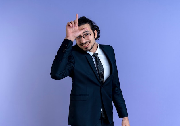 Business man in black suit and glasses looking to the front with loser sign over head smiling cheerfully standing over blue wall