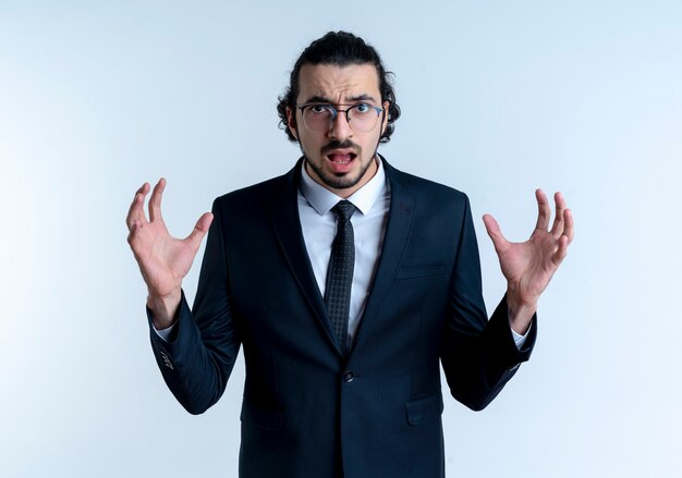 Business man in black suit and glasses looking to the front with raised arms with aggressive expression standing over white wall