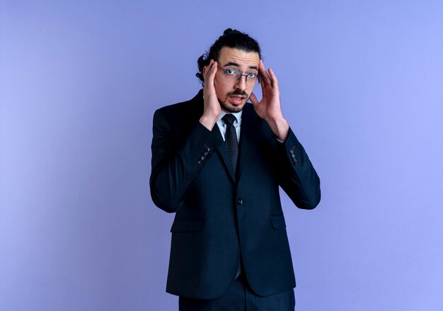 Business man in black suit and glasses looking to the front worried touching temples standing over blue wall