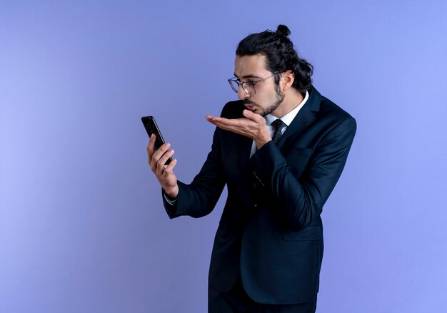 Business man in black suit and glasses looking at his smartphone screen on video call blowing a kiss standing over blue wall