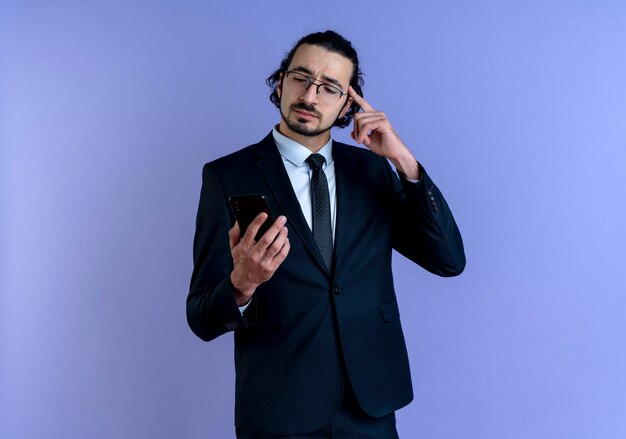 Деловой человек в черном костюме и очках, глядя на экран своего смартфона, указывая пальцем на висок, сосредоточился на задаче, стоящей над синей стеной