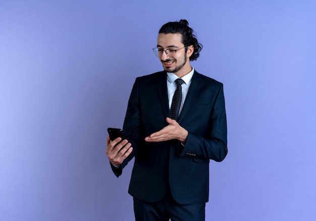 Free photo business man in black suit and glasses looking at screen of his smartphone smiling standing over blue wall