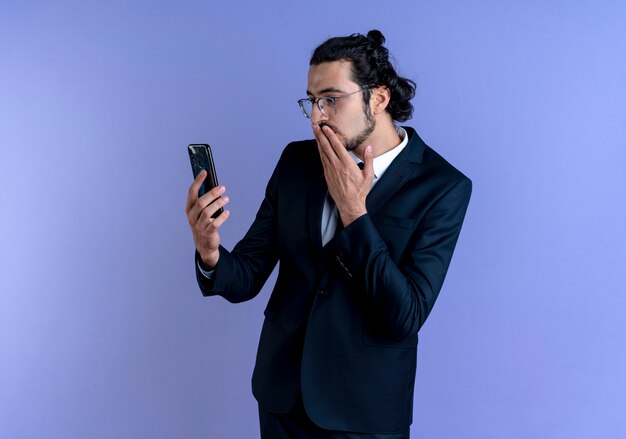 Деловой человек в черном костюме и очках, глядя на экран своего смартфона, удивлен и смущен, стоя у синей стены