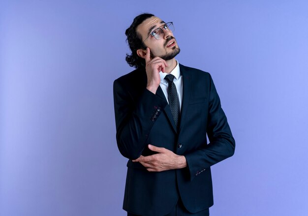 Business man in black suit and glasses looking up puzzled standing over blue wall