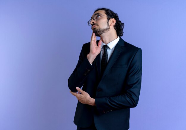 Business man in black suit and glasses looking up puzzled thinking standing over blue wall