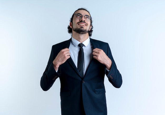 Business man in black suit and glasses looking up self-satisfied fixing his suit standing over white wall