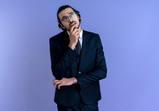 Business man in black suit and glasses looking up with hand on chin puzzled standing over blue wall