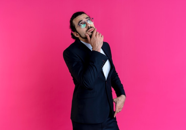 Business man in black suit and glasses looking up with hand on chin puzzled standing over pink wall