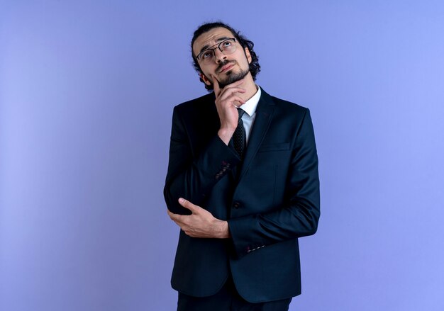 Business man in black suit and glasses looking up with pensive expression on face standing over blue wall