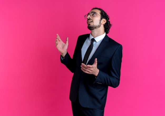 Uomo d'affari in abito nero e occhiali che osserva in su con la mano alzata confusa in piedi sopra il muro rosa