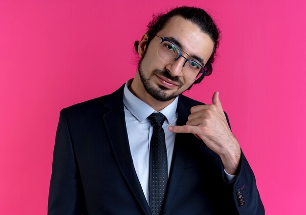 Business man in black suit and glasses making call me gesture with hand smiling confident standing over pink wall