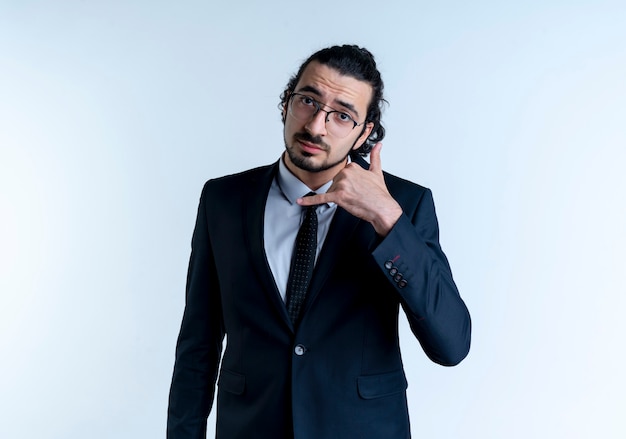 Business man in black suit and glasses making call me gesture with hand standing over white wall