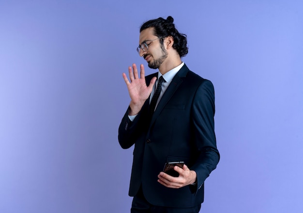 Business man in black suit and glasses making defense gesture agains his smartphone with disgusted expression standing over blue wall