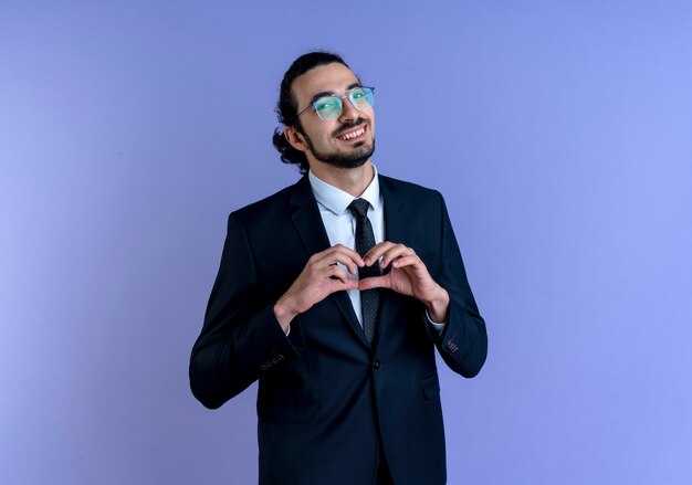 Business man in black suit and glasses making heart gesture with fingers over his chest looking to the front with smile standing over blue wall
