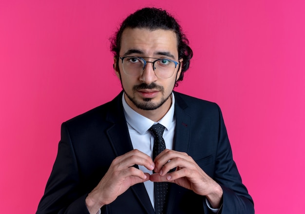Uomo d'affari in abito nero e occhiali che fanno il gesto del cuore con le dita che guardano in avanti con espressione fiduciosa in piedi sul muro rosa