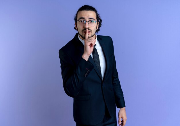 Business man in black suit and glasses making silence gesture with finger on lips standing over blue wall