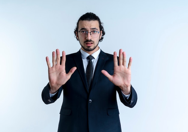 Business man in black suit and glasses making stop sign with both hand looking to the front with serious face standing over white wall