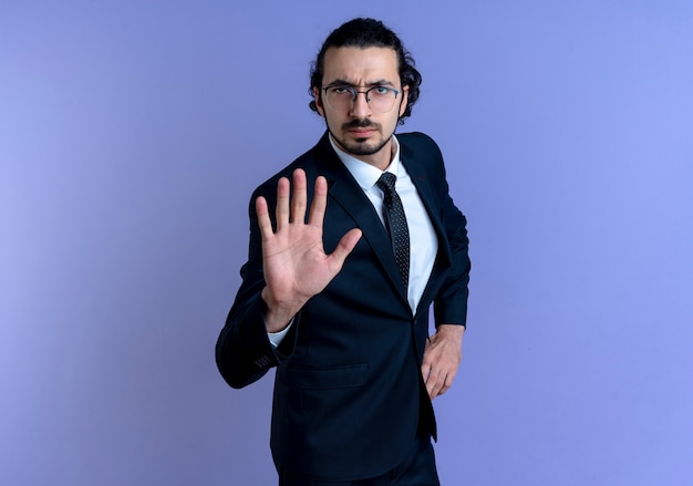 Business man in black suit and glasses making stop sign with hand looking to the front with serious face standing over blue wall