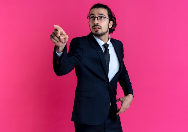 Деловой человек в черном костюме и очках, указывая пальцем в сторону, выглядит удивленным, стоя над розовой стеной