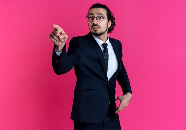 Business man in black suit and glasses pointing with finger to the side looking surprised standing over pink wall