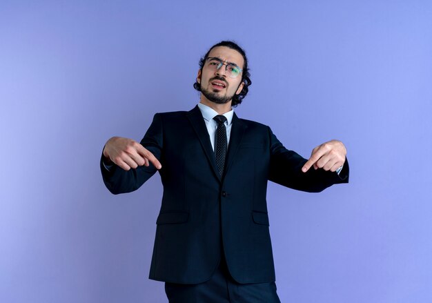 Деловой человек в черном костюме и очках, указывая пальцами на себя и недовольный, стоит над синей стеной