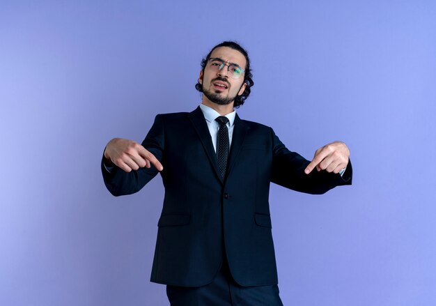 Business man in black suit and glasses pointing with fingers to himself looking displeased standing over blue wall