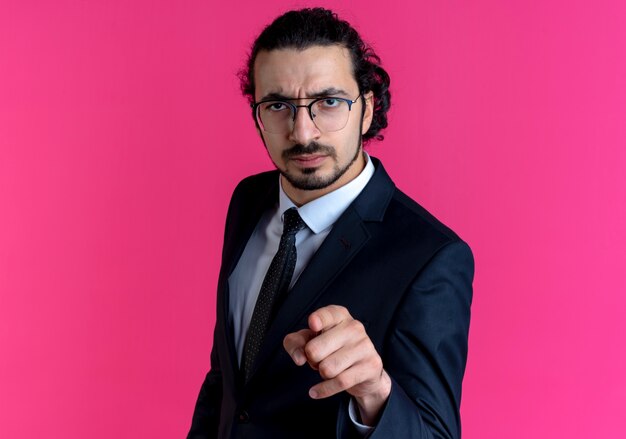 Business man in black suit and glasses pointing with index finger to the front with serious face standing over pink wall