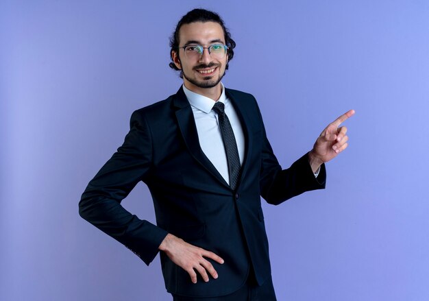 Business man in black suit and glasses pointing with index finger to the side smiling cheerfully standing over blue wall