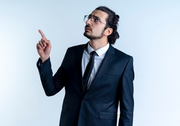 Business man in black suit and glasses pointing with index finger up looking confident standing over white wall