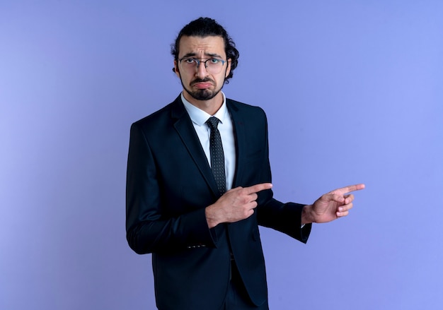 Business man in black suit and glasses pointing with index fingers to the side looking confused with sad expression standing over blue wall