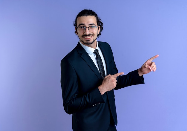 Business man in black suit and glasses pointing with index fingers to the side smiling cheerfully standing over blue wall