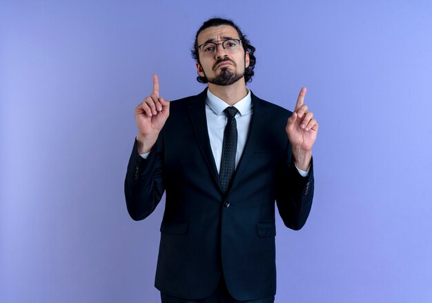 Business man in black suit and glasses pointing with index fingers up looking to the front with sad expression standing over blue wall