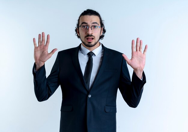 Business man in black suit and glasses raising arms in surrender looking to the front with fear expression standing over white wall